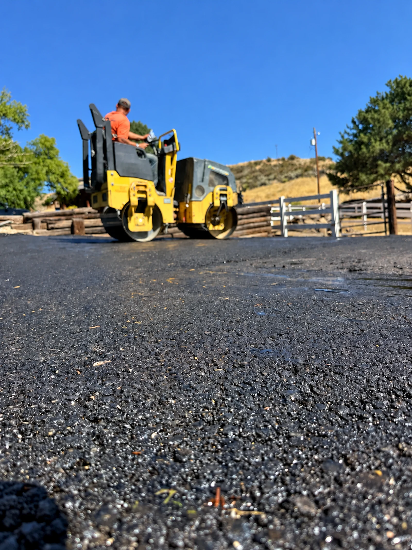 A worker drives a yellow roller across dark pavement.